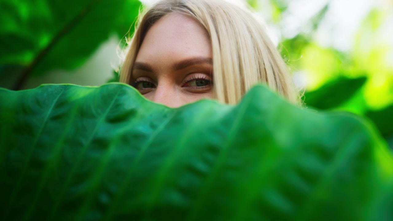Primo piano di una giovane, attraente donna caucasica bionda che sposta una foglia tropicale verde in un ambiente tropicale caldo, rivelando occhi bellissimi in modo giocoso. Retroilluminata dal sole. Rallentatore.
