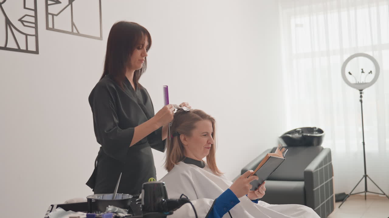 Parrucchiere che acconcia i capelli di una donna seduta su una sedia in un salone, parrucchiere professionista che pettina e asciuga i capelli di media lunghezza, cliente che scorre sullo smartphone sotto la luce naturale soffusa, ring light e moderno studio minimal, vassoio