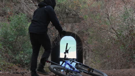 man-walks-towards-a-bicycle,-picks-it-up,-and-leaves,-while-tunnel-leading-to-wild-sea-centres-frame