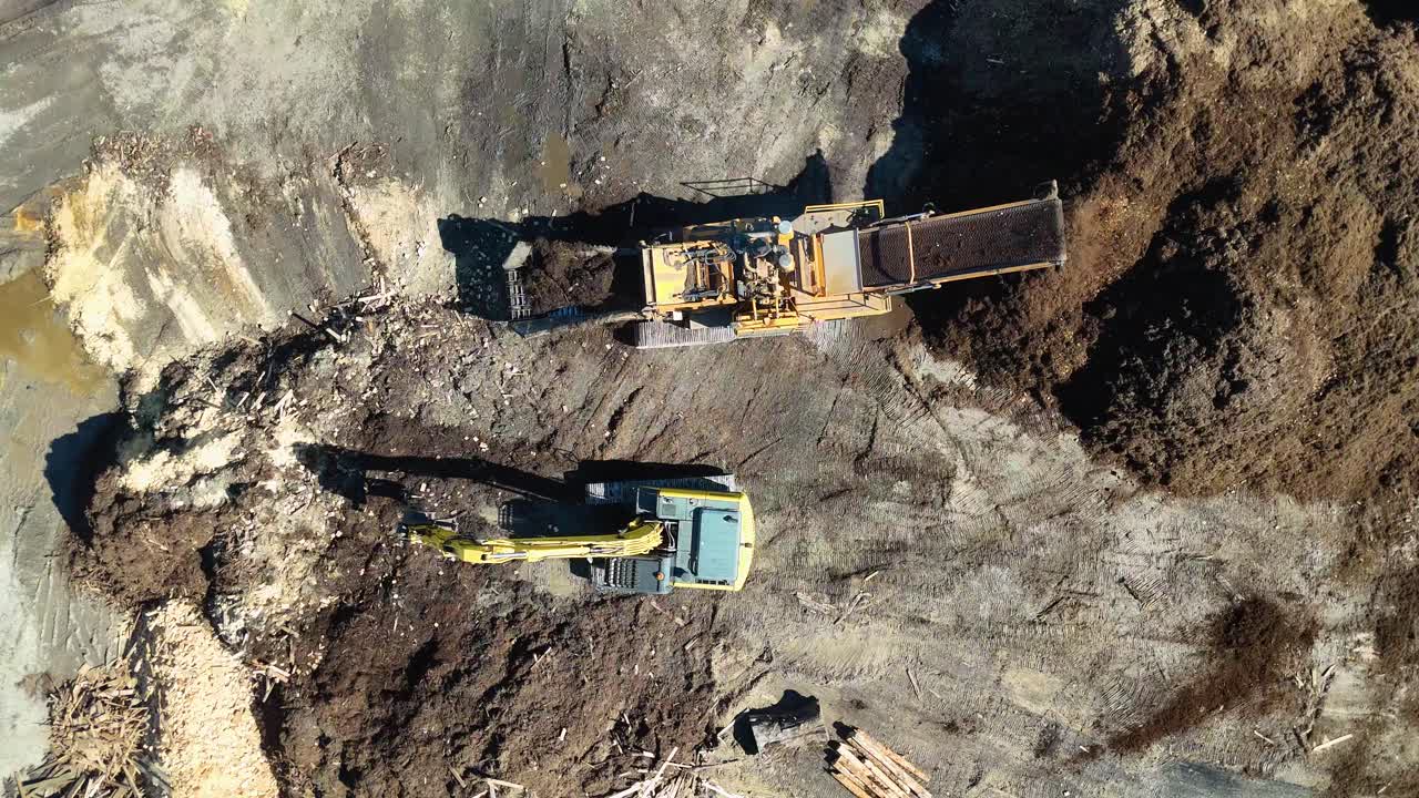 Drone footage captures an excavator moving mulch in a dirt processing area under bright sunlight