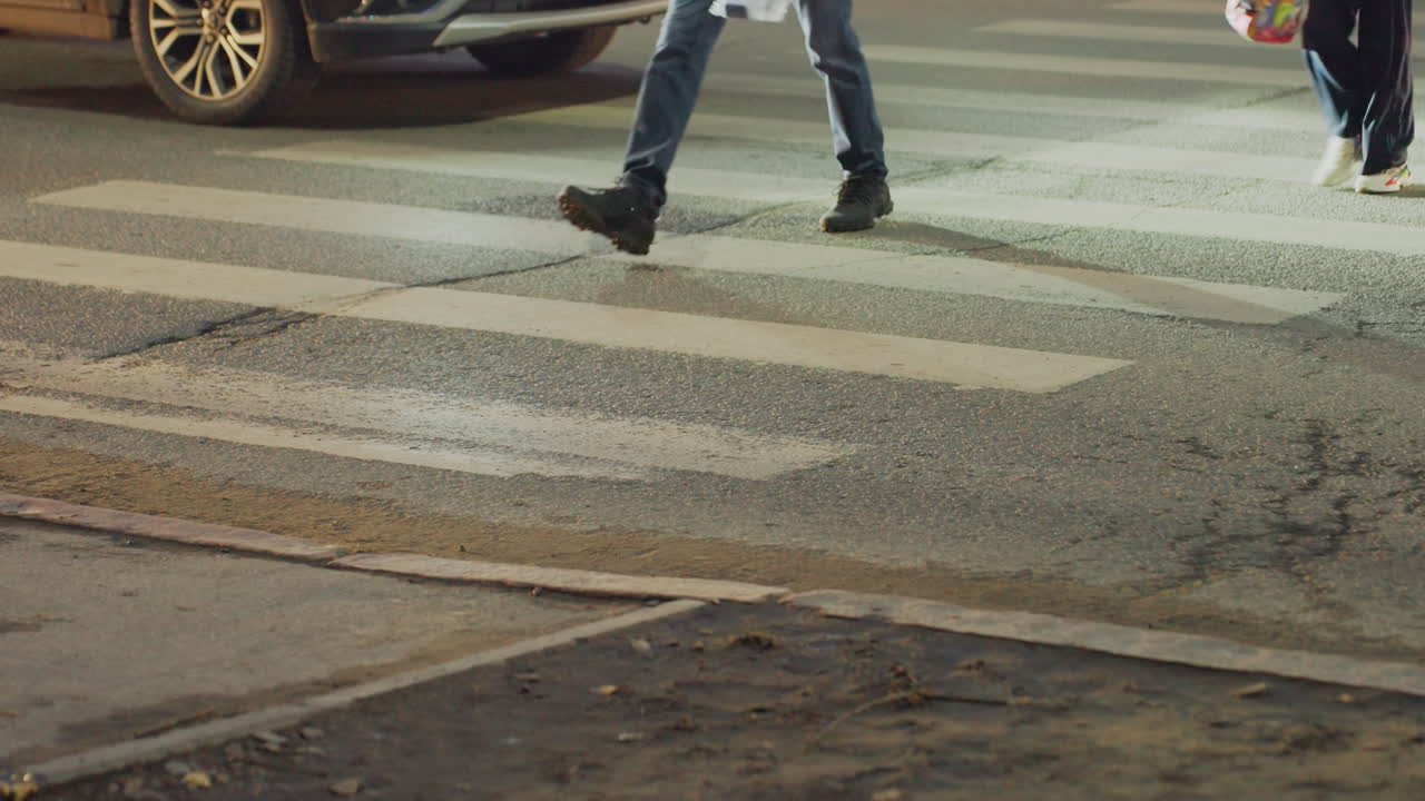 Legs of pedestrians crossing white striped crosswalk on city street as vehicle approaches from side during evening time, with asphalt reflecting soft light and urban pavement details visible