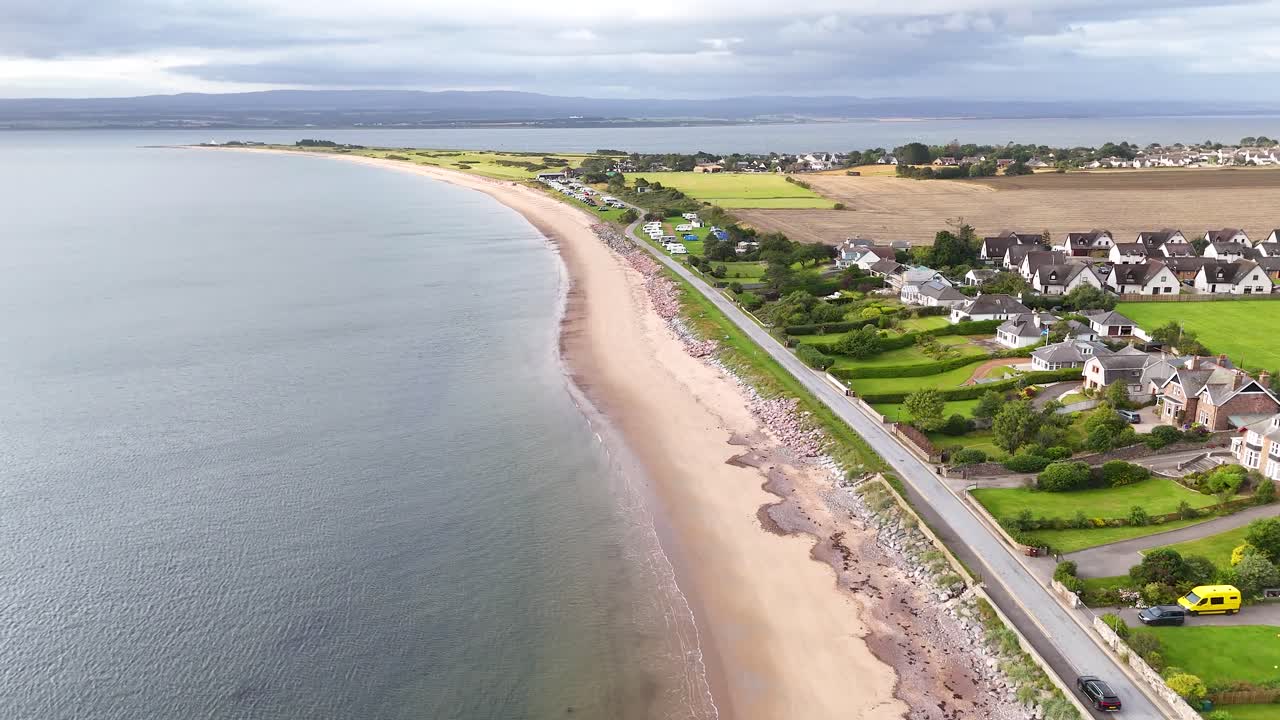Drone footage glides above a sandy shoreline, coastal homes, and green fields in Rosemarkie, Scotland, under soft daylight with steady camera movement