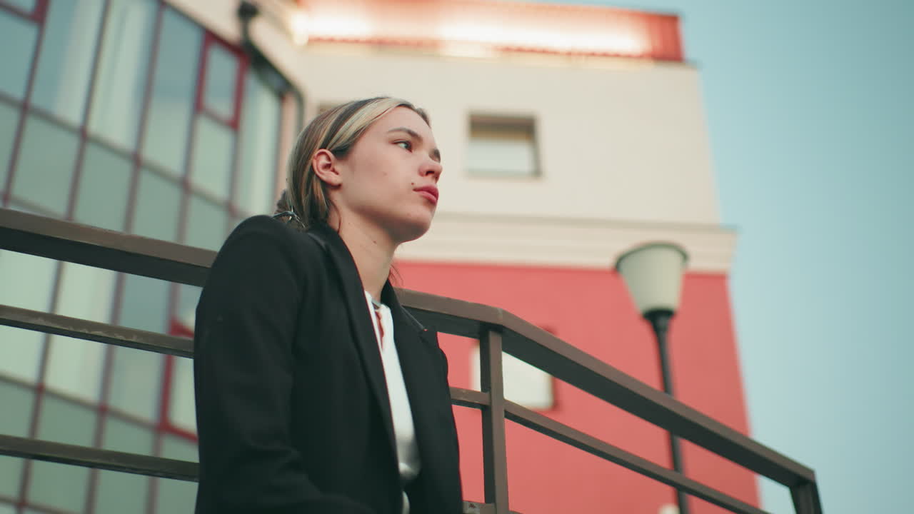 Disturbed lady in black blazer and white top waiting for delivery seated on fence, looking at phone with modern building in background and light blinking on roof
