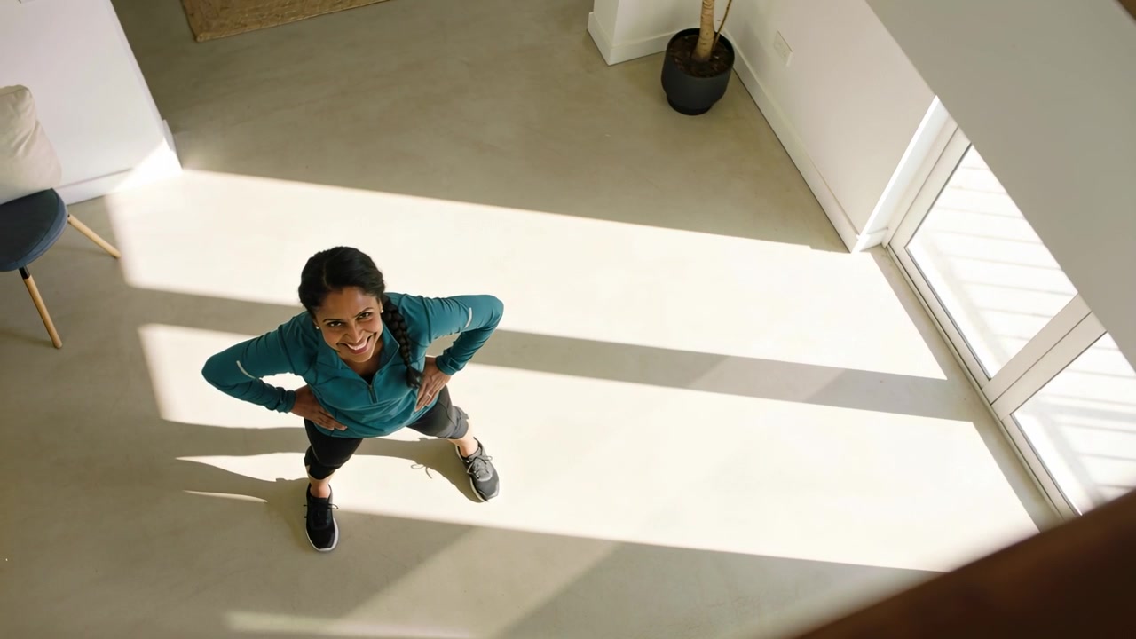Standing woman in teal zip-up top shifting feet, looking up at overhead camera, preparing exercise