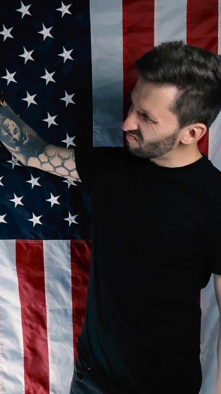 Uomo tatuato con una mascherina protettiva nera per il viso (COVID o sanitaria) si toglie la mascherina e la getta via con disgusto mentre è in piedi davanti a una bandiera verticale degli Stati Uniti d'America. L'uomo è stufo.
