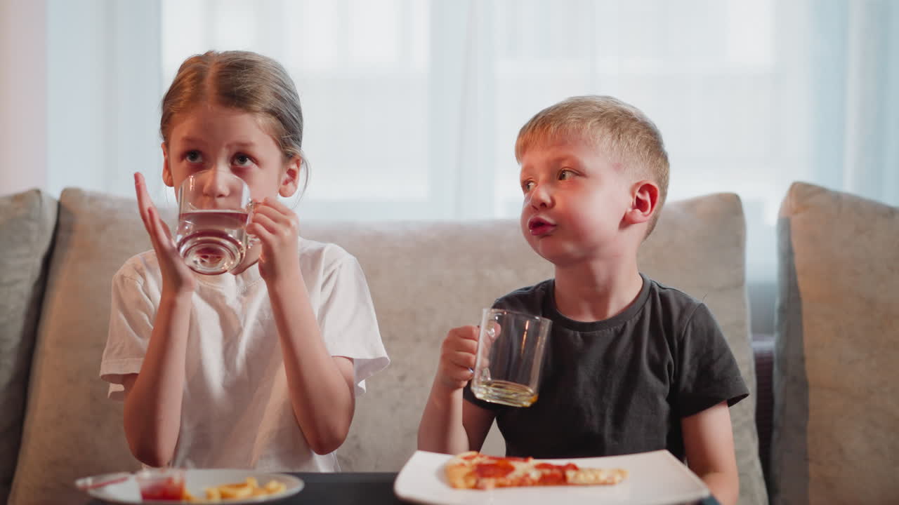 brother and sister sitting together at dining table with boy picking up glass of juice while sister drinks water boy looks unhappy covering face as they share conversation