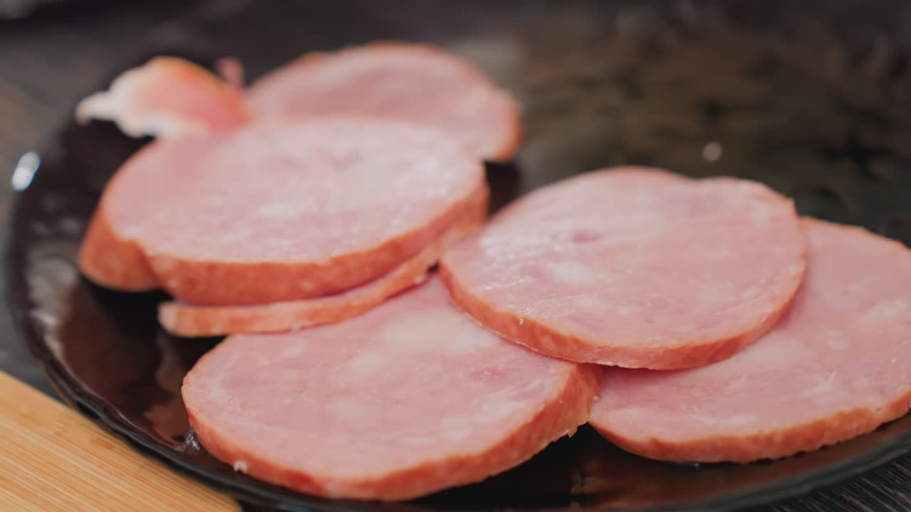 close up hand view of fair skin person arranging round pink sausage slices on glossy black plate, meat pieces overlapping in neat stack, soft lighting and shallow focus