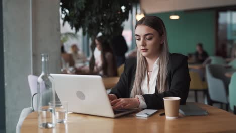 beautiful, stylish young woman sits in a cafe in business attire, opens her laptop, and starts working with a smile