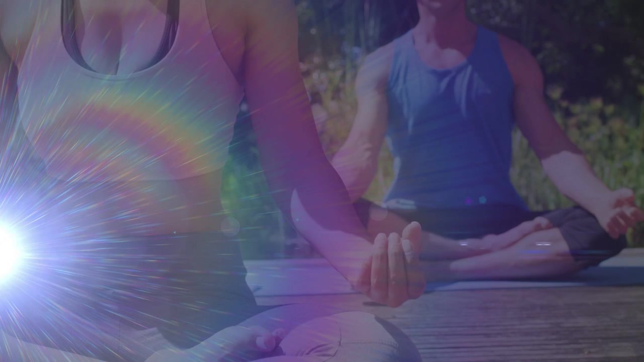 Homem abaixando as mãos de uma posição de oração enquanto mulher forma um mudra, praticando yoga no deque com um brilho.