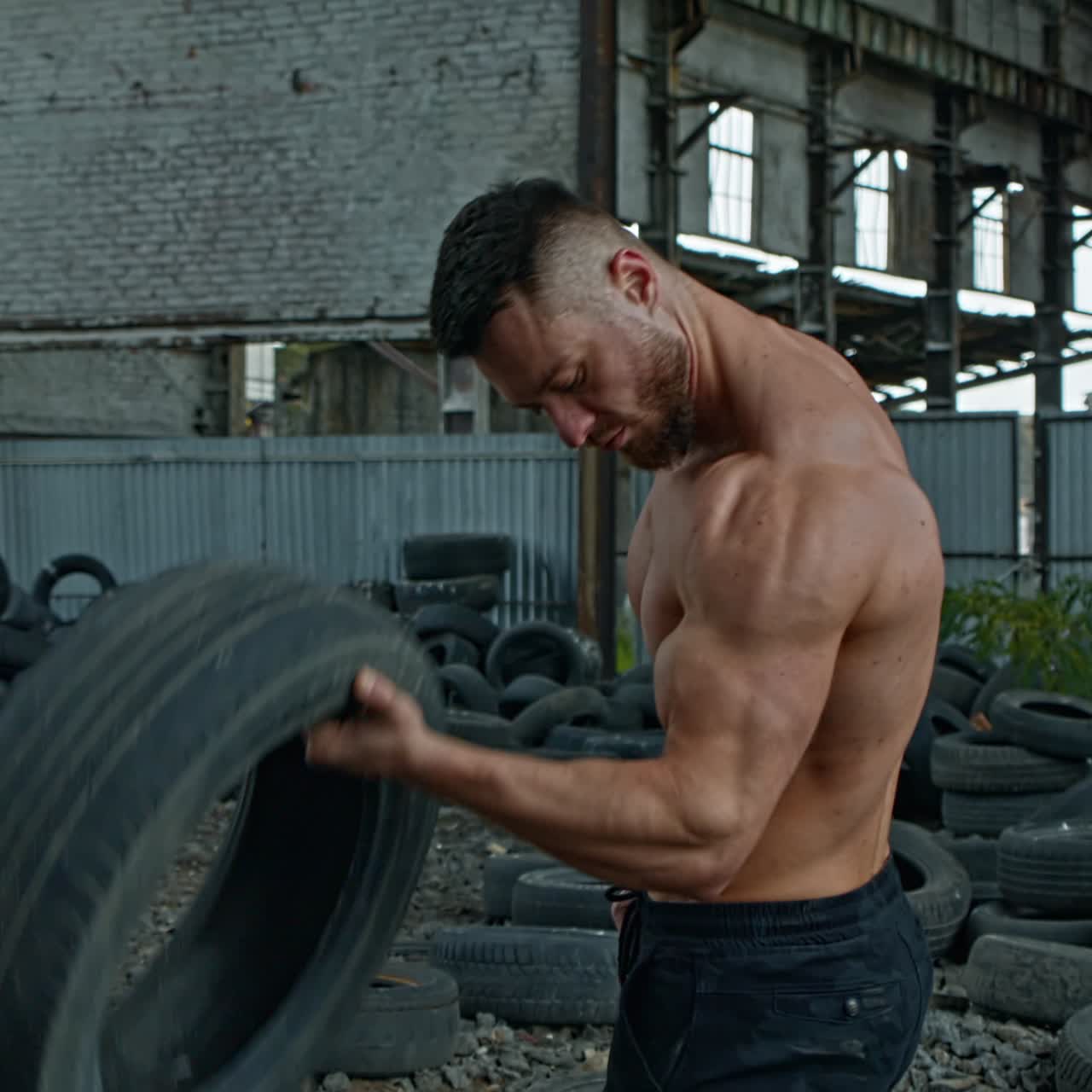 Male athlete with tires in abandoned place. Strong young man training his arm muscles with car tyre during an intense training session on ruined building background.