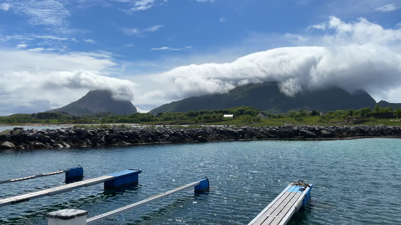 밝고 화창한 날 작은 정박지의 아름다운 패닝 샷을 배경으로 구름으로 뒤덮인 산과 바람이 물결을 일으키고 있습니다.
