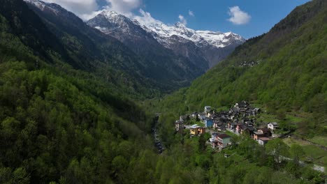 Hermosa-Toma-En-órbita-Aérea-De-Un-Pequeño-Pueblo-Metido-En-Un-Verde-Profundo-Arbolado-Valle-Cortado-Por-El-Río-Con-Espectaculares-Montañas-Gigantes-Cubiertas-De-Nieve-Que-Se-Elevan-Sobre-El-Valle