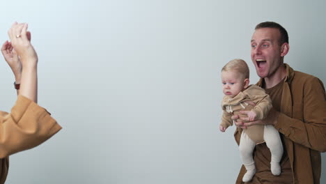 man holds a cute blond baby boy. woman touches her child tickling him and smiling to a kid. white backdrop.