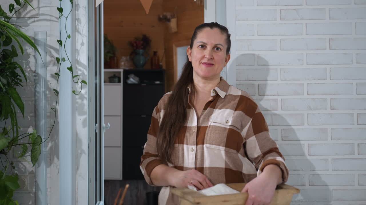 Portrait of gardener smiling while carrying basket with fresh plant clippings and gardening tools stands before open door brick wall indoor cozy greenhouse morning light warming friendly mood