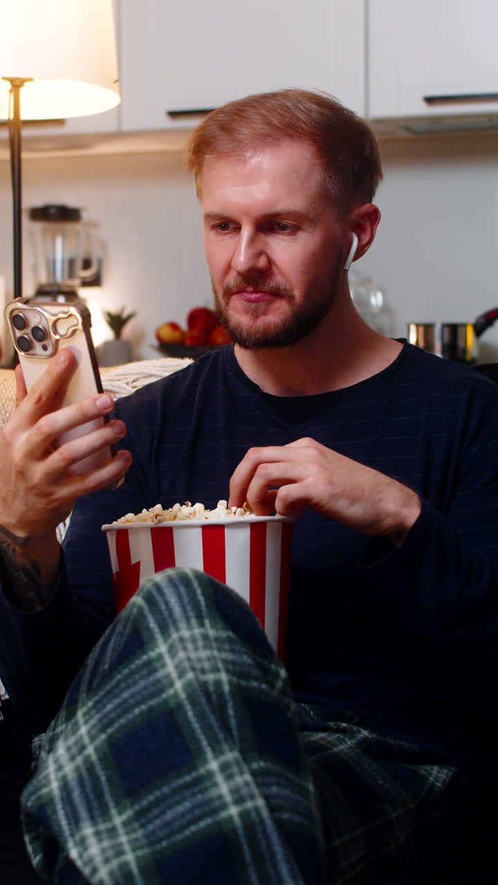 Giovane uomo sul divano guarda serie in streaming sullo smartphone, ride a crepapelle, mangia popcorn e si rilassa a casa la sera.