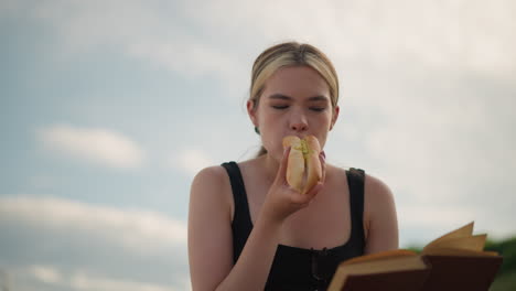 woman seated outdoors holding book in one hand while eating sausage roll and reading, blurred background features a lamp post and people walking in the distance