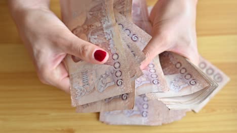 high angle view of woman with red fingernails counting thai baht banknotes on wooden table.