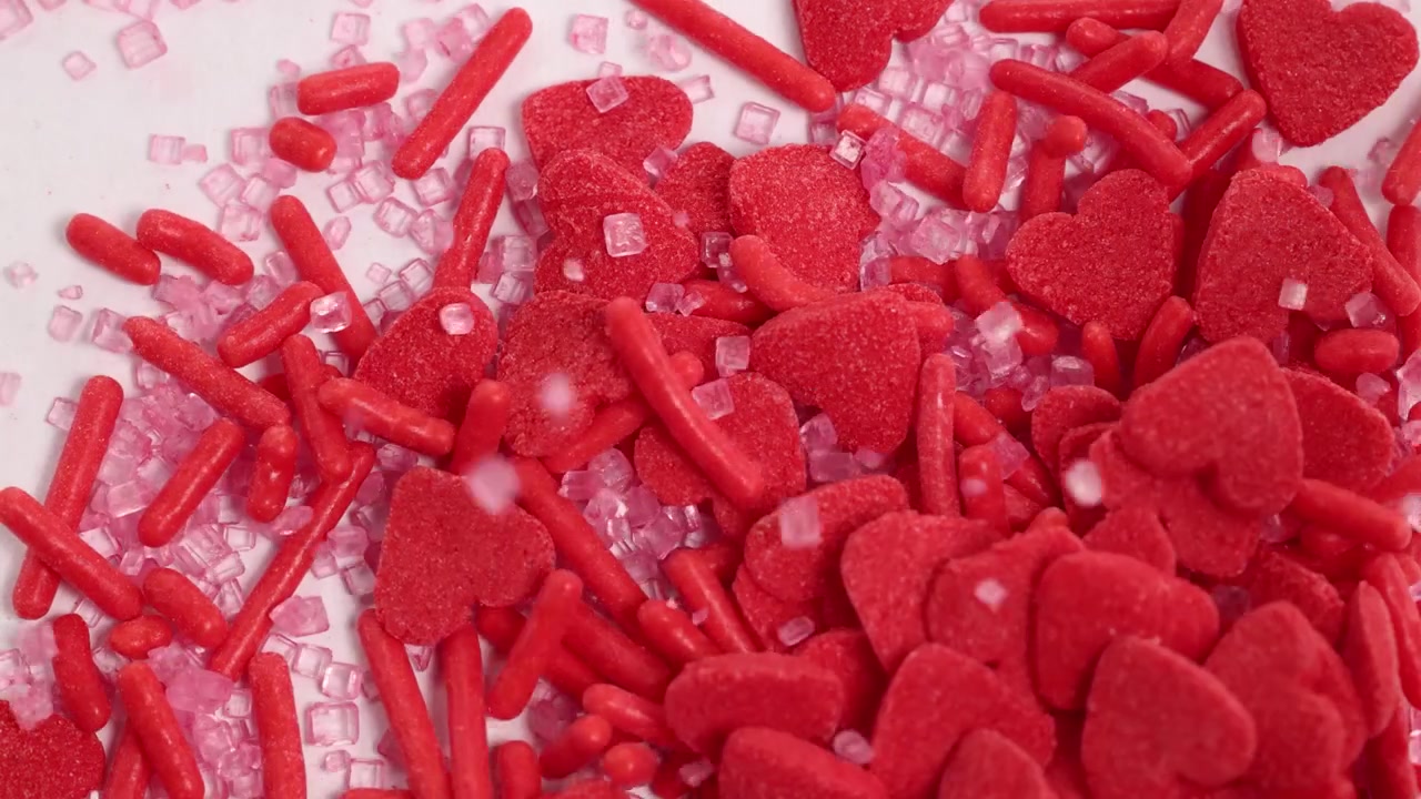 Red heart-shaped candies and sprinkles cascade onto a white surface, creating a vibrant, sweet composition