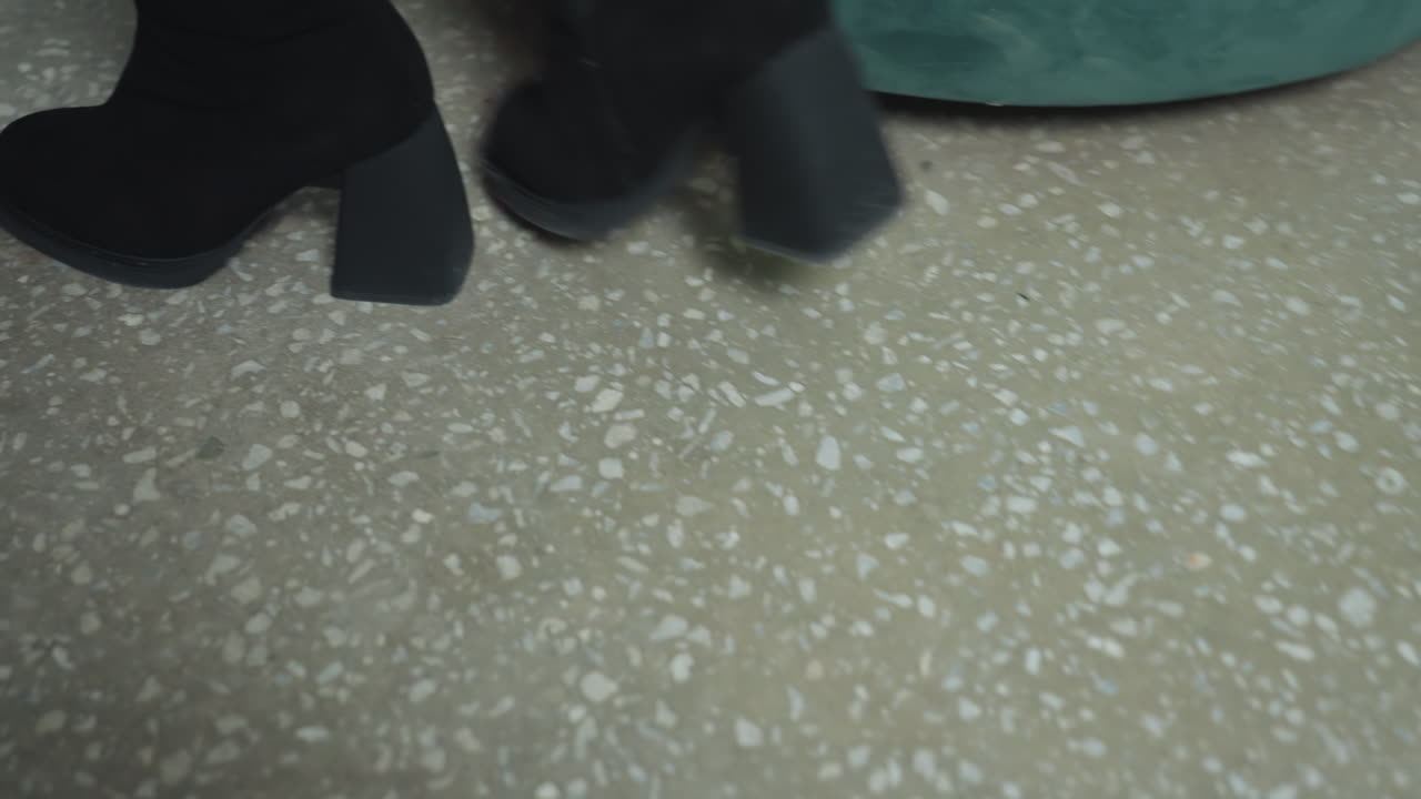 Close up leg view of woman in black heel boots walking on terrazzo floor toward green couch, stylish confident motion captured from low angle emphasizing footwear design