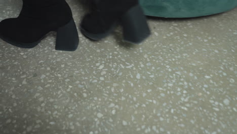 close up leg view of woman in black heel boots walking on terrazzo floor toward green couch, stylish confident motion captured from low angle emphasizing footwear design