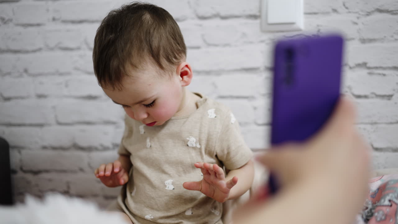 Adult's hand showing a phone to a little toddler. Baby looks at gadget with interest. White wall at backdrop.