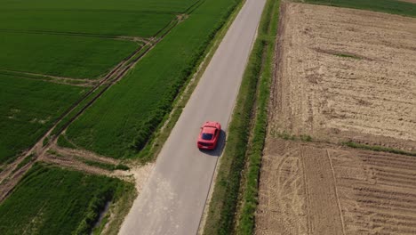 Filmische-Drohnenaufnahme-Eines-Orangefarbenen-Ford-Mustang-GT,-Der-Eine-Gerade-Straße-Entlang-Fährt,-Umgeben-Von-Grünen-Feldern