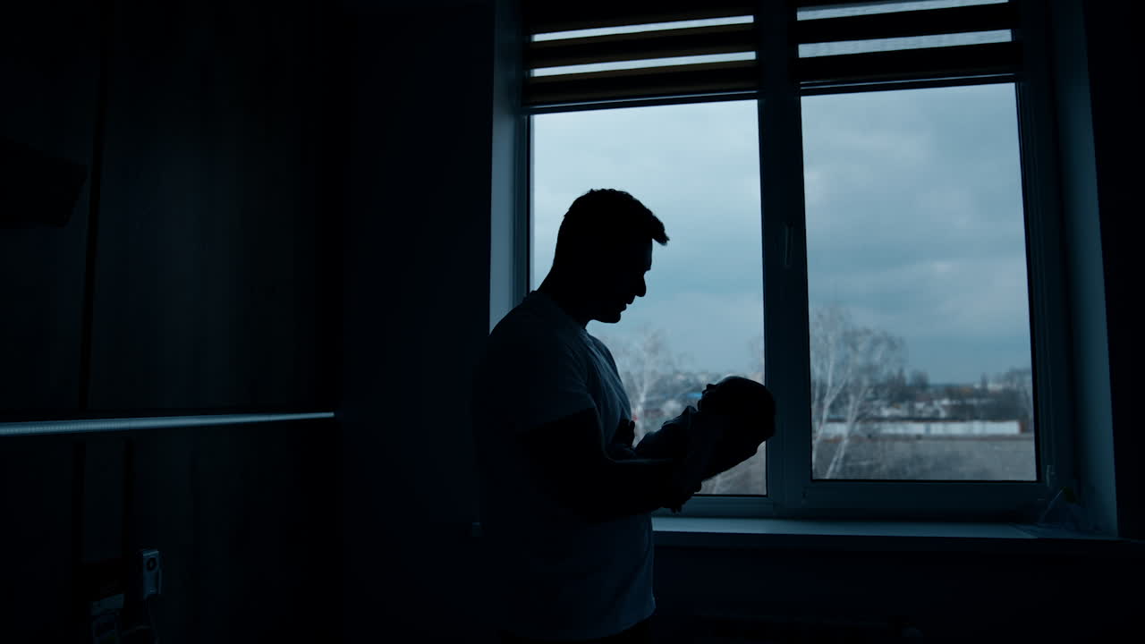 Silhouette of a man holding a baby in hands in front of him. Father with a child standing at the window.