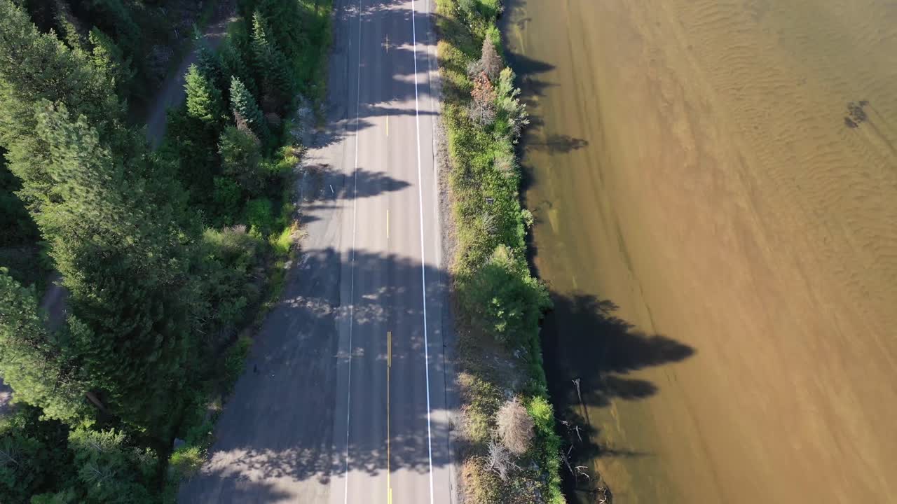 Drone tilts up with an increasing push in over a winding road that is lined with forest trees and runs parallel to a wide glossy river.