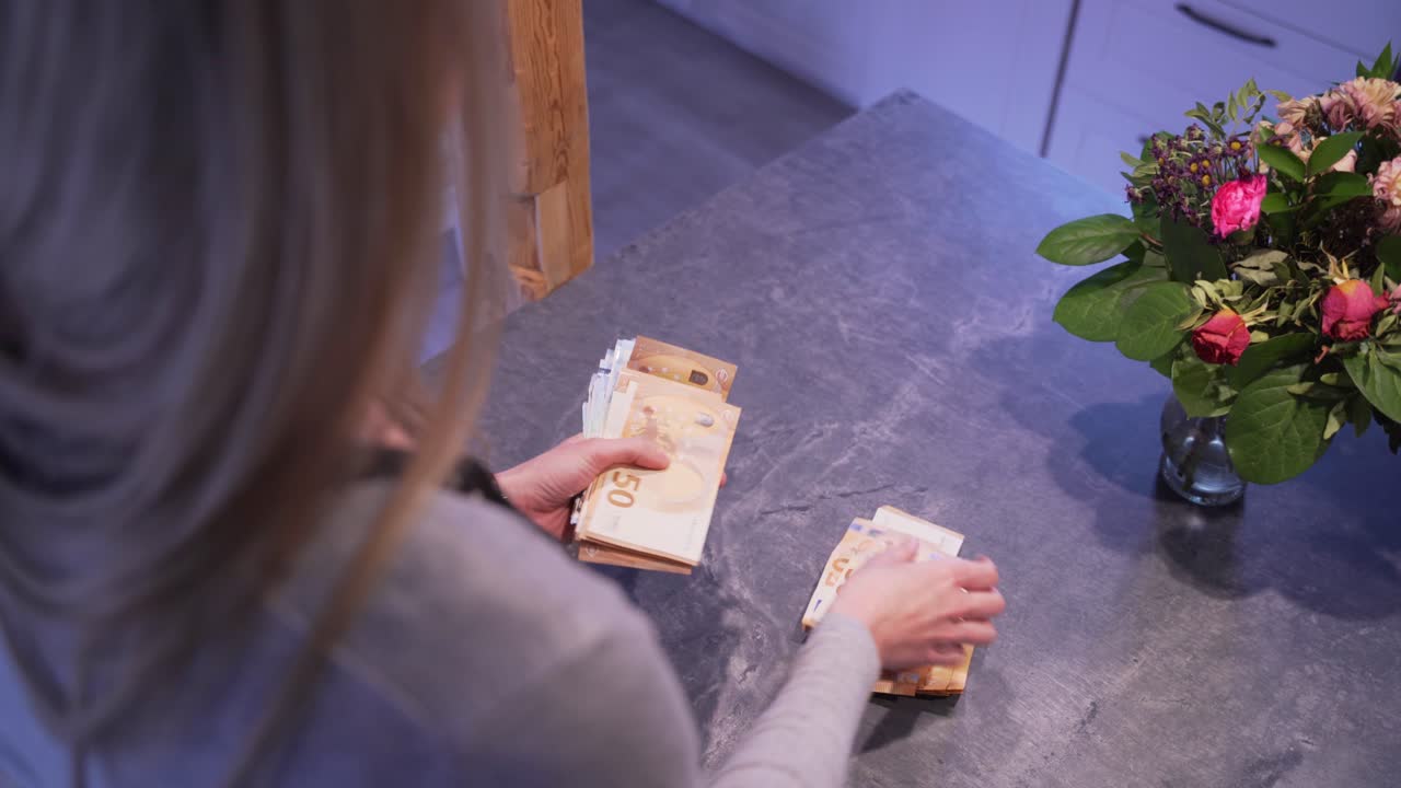 A blonde girl is counting 2000€ on a black table. There are two 100€ bills, some 50s and some 20s. There are beautiful flowers standing on the table.