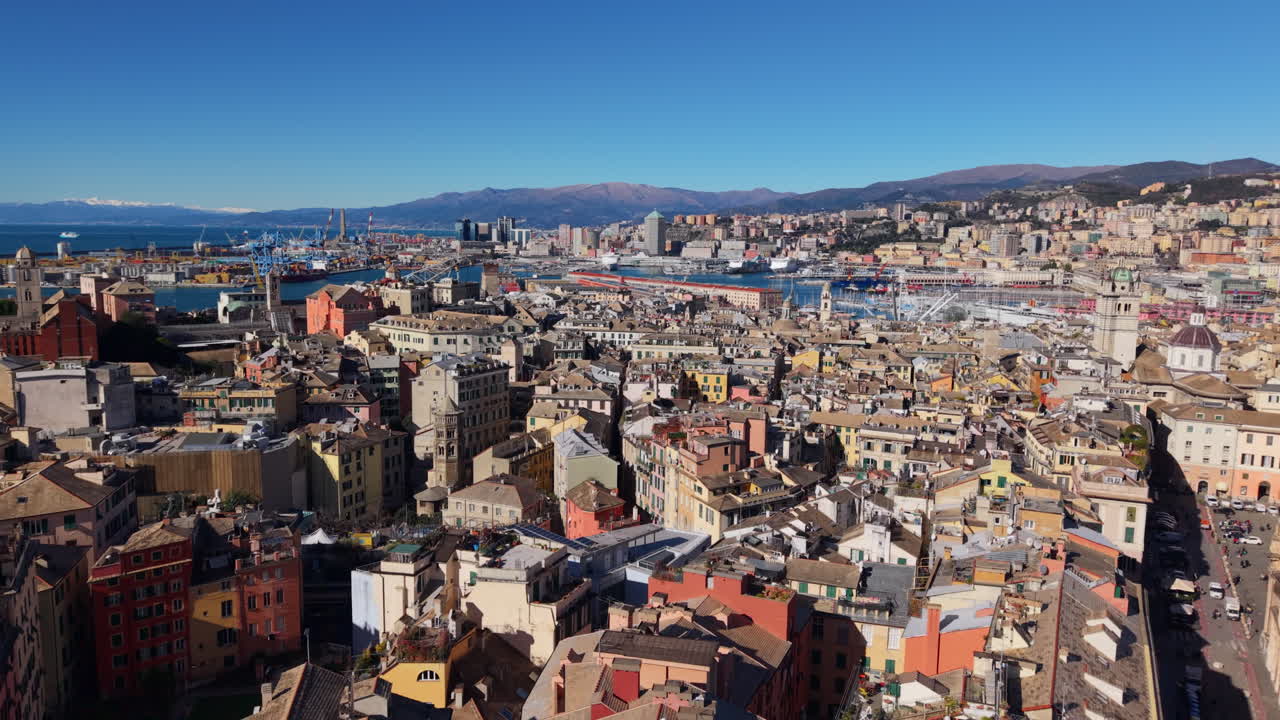 Drone che vola in avanti sopra il centro storico di Genova in Liguria, Italia, dirigendosi verso il porto commerciale con gru portacontainer e montagne sotto un cielo azzurro brillante.