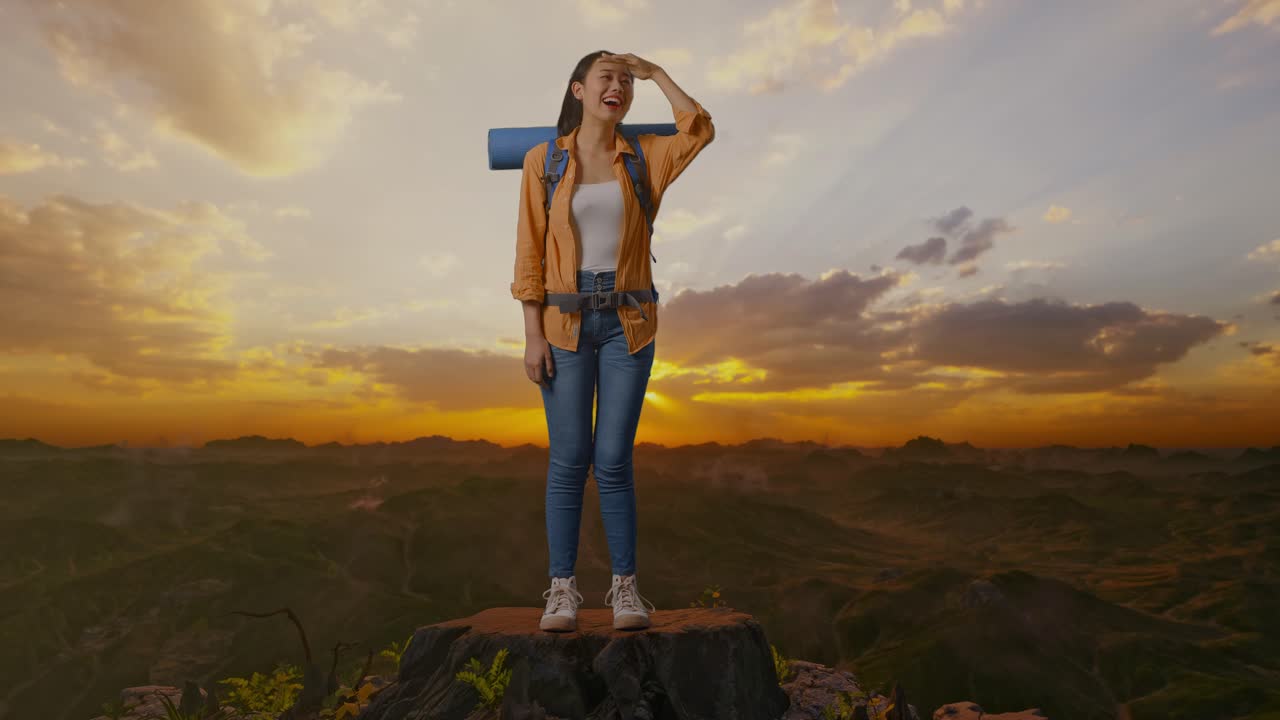 Escursionista asiatica a figura intera con zaino da trekking, con la mano sulla fronte, che sorride e guarda lontano mentre si trova in cima alla montagna al tramonto