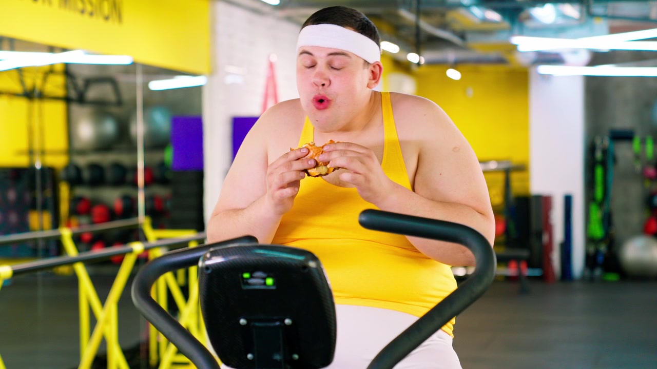 Uomo in sovrappeso che mangia un hamburger su una cyclette in palestra