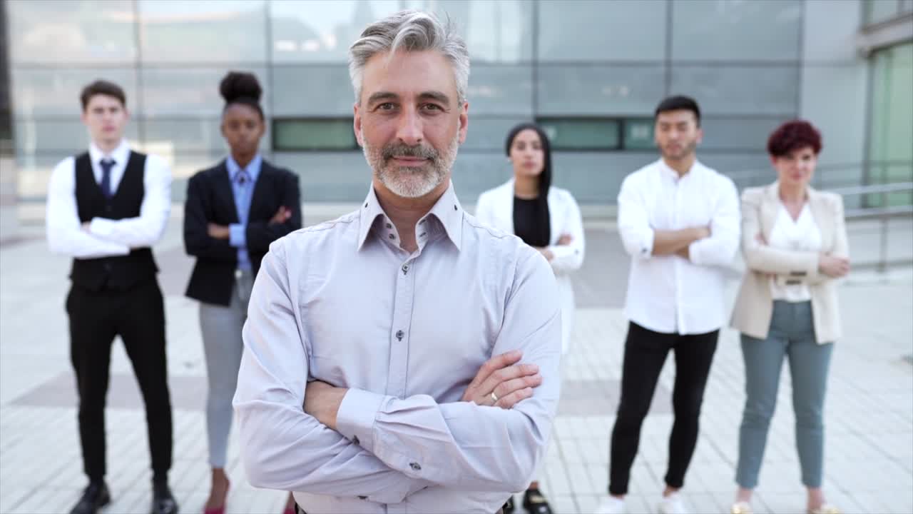 video de un empresario con los brazos cruzados, sonriendo feliz, en el fondo sus compañeros de trabajo en una reunión al aire libre