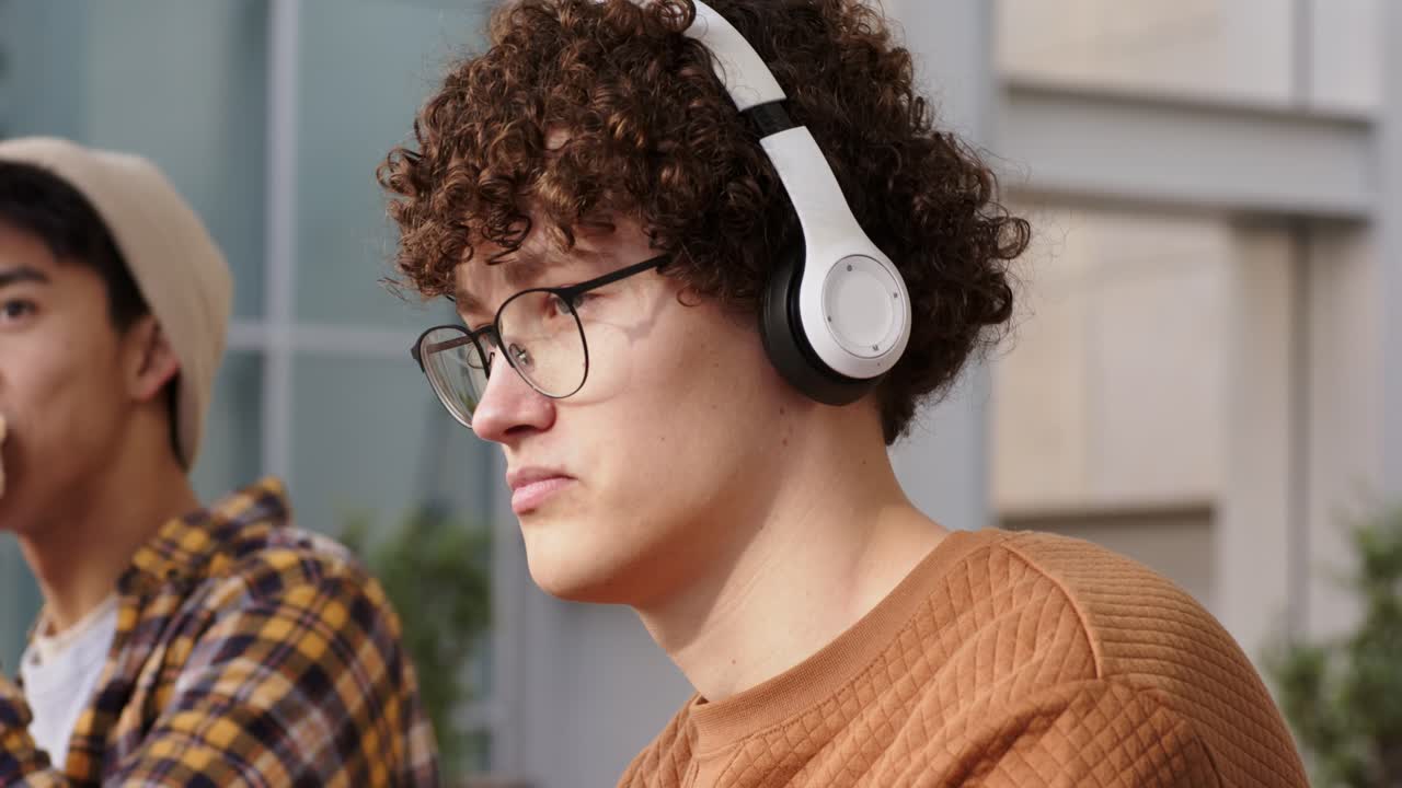 Diversos amigos do sexo masculino compartilhando um banco enquanto um deles alcança, ajusta e ouve algo depois de ver fones de ouvido