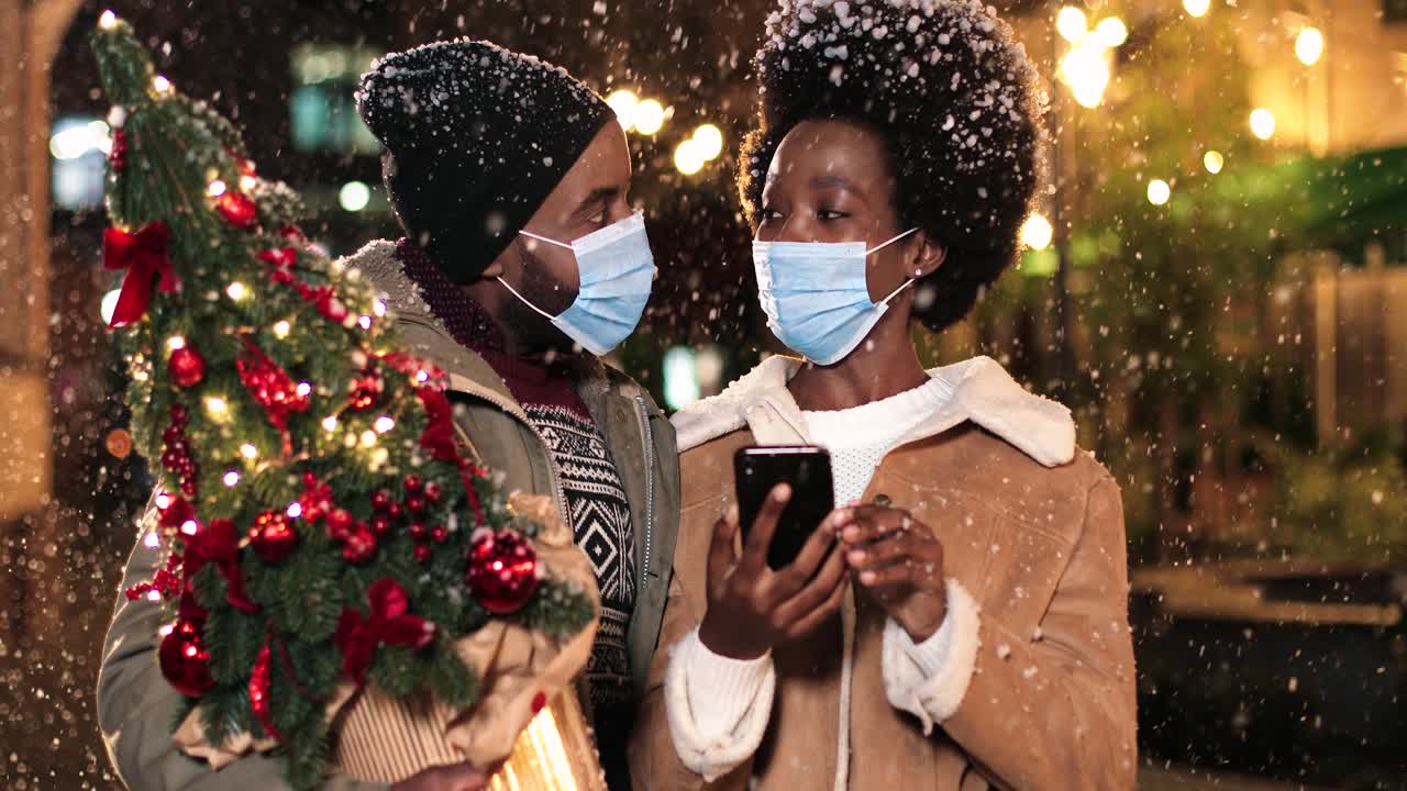 Close-up view of joyful African American couple wearing facial masks talking and watching something on the phone while it¬¥s snowing on the street in Christmas