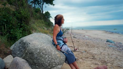 old woman sitting on a huge rock by the beach in karkle, lithuania - wide shot