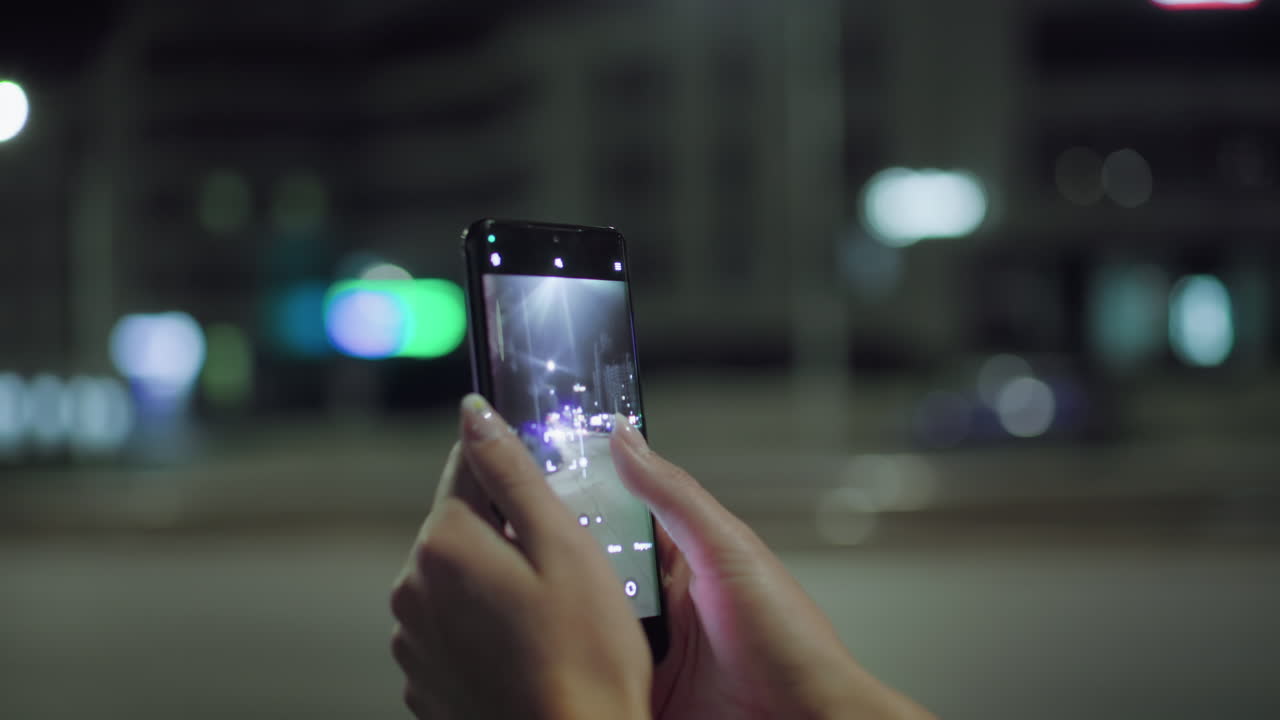 Close up of woman capturing night photo with smartphone on roadside, colorful blurred city lights in background, cars passing by under bright streetlights