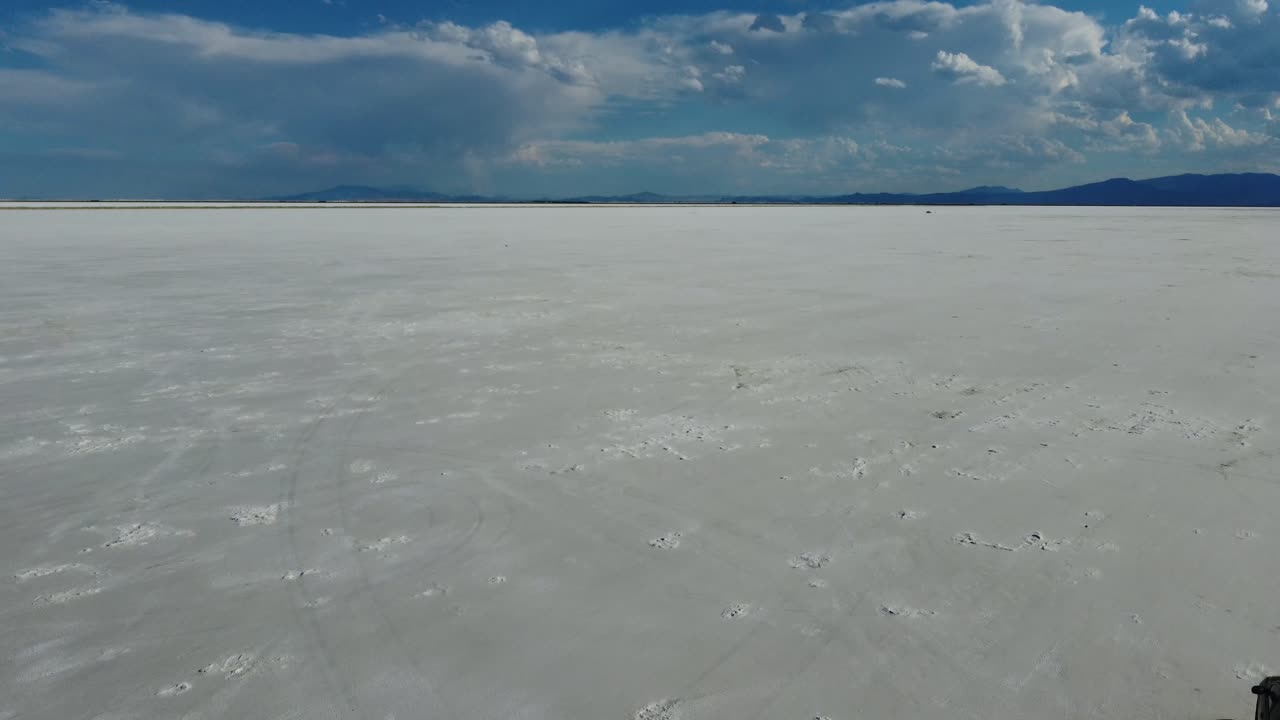 Car driving to the right of the camera as the drone flies by. Filmed at the Bonneville Salt Flats in Utah during summer. Contrasted by the blue sky and white sand. 4K drone footage.