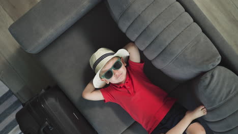 adorable niño en sombrero de verano se relaja en el sofá cerca de la maleta