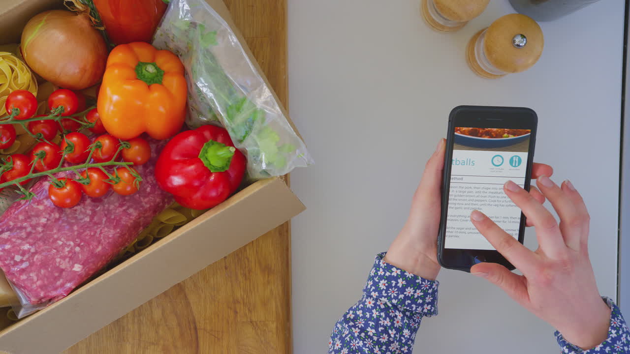toma aérea de una mujer en la cocina con ingredientes frescos mirando una receta en línea en un teléfono móvil - tomada en cámara lenta