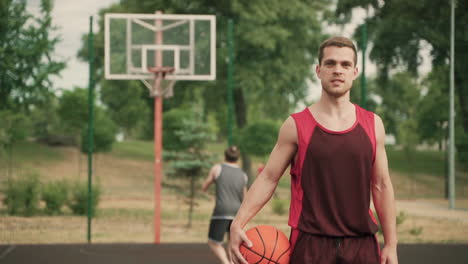 in foreground, an handsome basketball player with ball, breathing heavily after a good productive training session, while looking at camera and smiling confidently