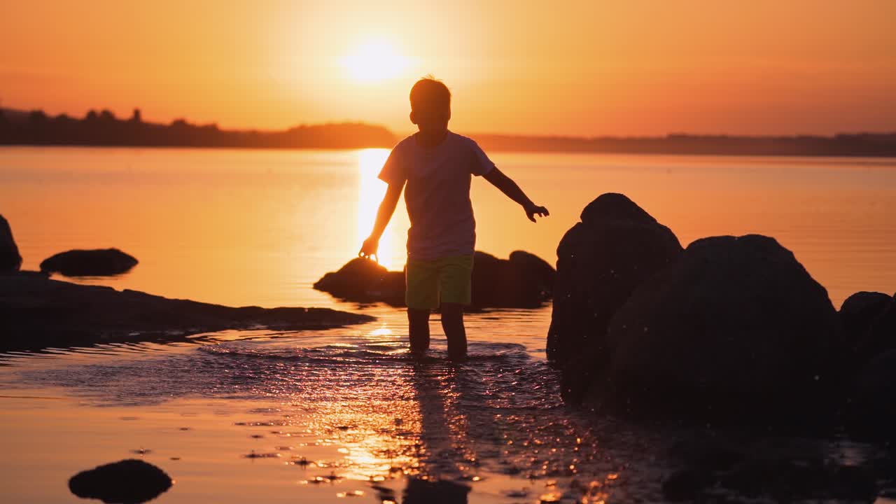 Boy walking in water at sunset. Silhouette of a child in clothes standing inside the river among stones. Kid splashing in the evening water. Slow motion.