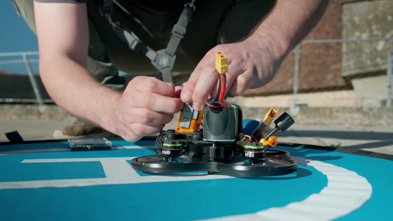 Hands of a pilot connecting a yellow battery plug to a racing drone placed on a blue landing pad during pre-flight preparation