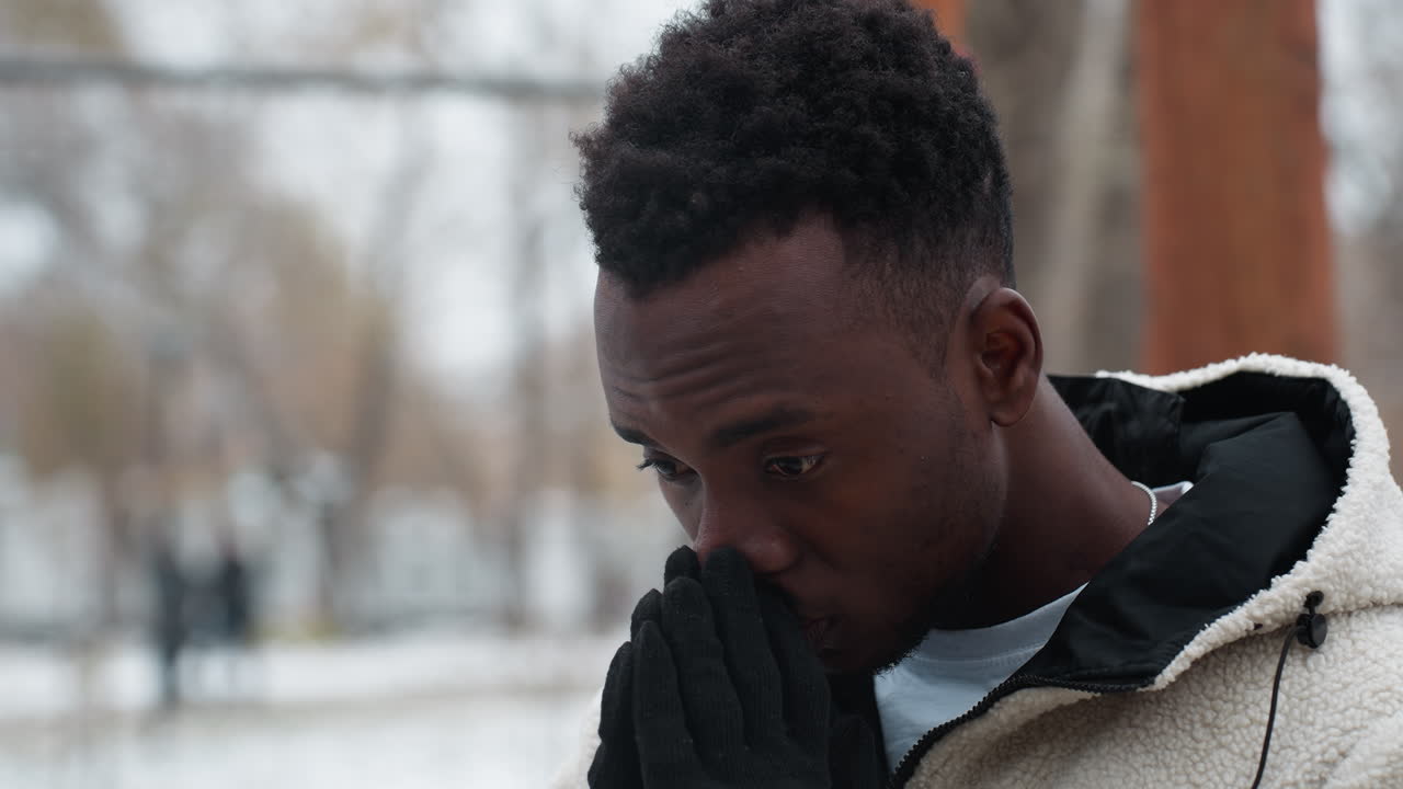 hombre pensativo con guantes negros y chaqueta blanca de invierno se encuentra al aire libre en clima frío, manos cubriendo la boca, fondo cubierto de nieve con árboles y edificios borrosos