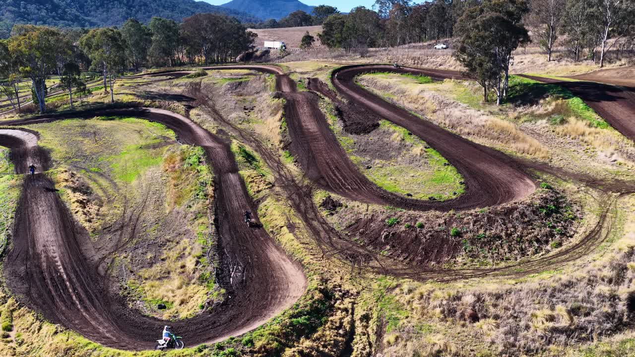 A motorbike rider navigates a winding dirt track, launching over a jump in a sunlit, rural Australian landscape, captured by a drone in wide aerial view