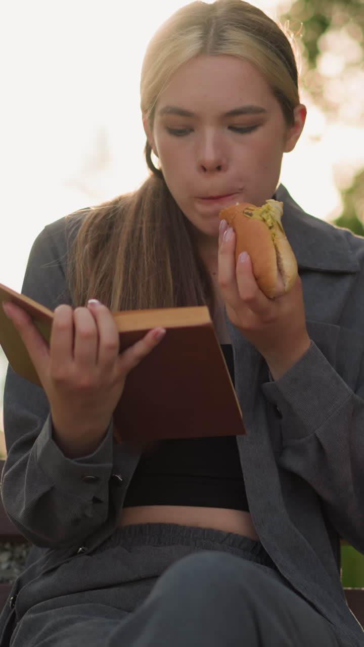 회색 을 입은 여자가 야외에서 책을 읽는 동안 간식을 먹고, 나무와 초록색으로 둘러싸인 공원 벤치에 앉아, 흐릿한 배경으로 부드러운 빛이 비추고 있습니다.