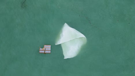 Large white piece of plastic sail floats in murky green water next to broken wooden block, drone