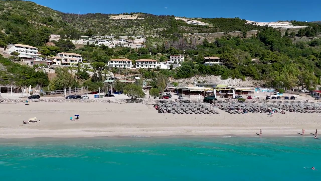 Drone voando da água turquesa em direção a um resort à beira-mar e uma encosta costeira. Destino litorâneo pitoresco com guarda-sóis, praia, mar cristalino e paisagem mediterrânea.