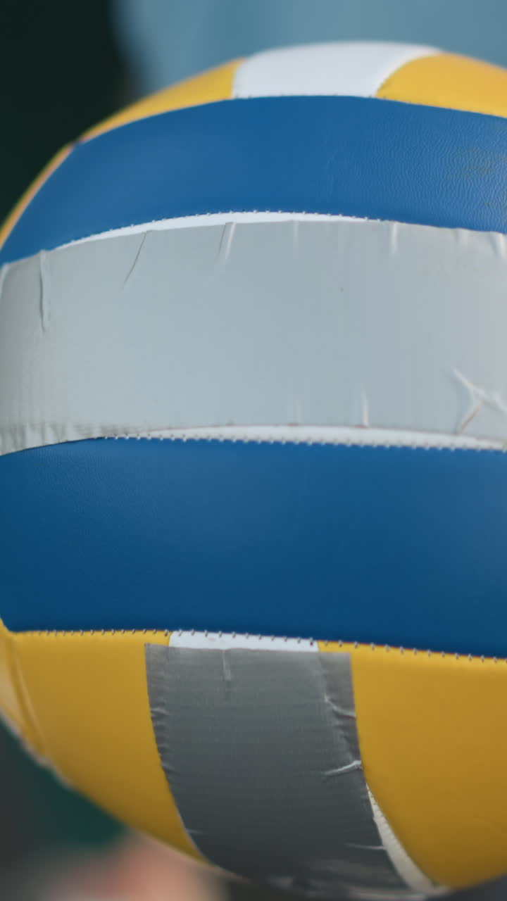 Primo piano di una pallavolo tenuta in mano da una donna, mentre un'altra persona con un maglione color vinaccia le mostra come servire. Entrambe sono concentrate sull'apprendimento e sulla pratica della tecnica all'aperto