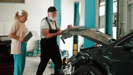 un viejo mecánico en el taller abre el capó de un coche destrozado y comienza una inspección y evaluación de los daños en presencia de un agente de seguros.